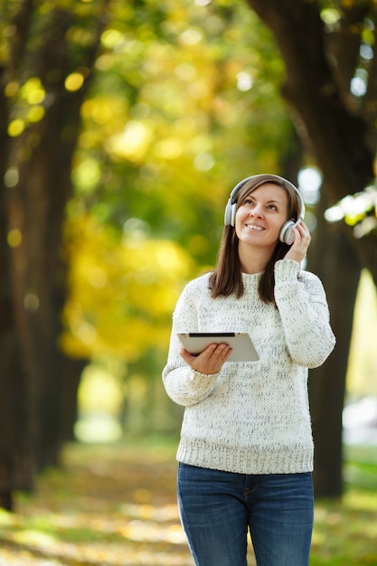 Die schöne fröhliche, fröhliche braunhaarige Frau im weißen Pullover mit einem Tablet, das an einem warmen Tag Musik in den weißen Kopfhörern im Herbstpark hört. Herbst in der Stadt.