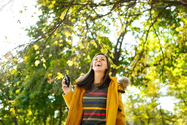 Die schöne fröhliche, fröhliche braunhaarige Frau in gelbem Mantel und gestreiftem Langarm, die an einem warmen Tag im Herbststadtpark mit dem Handy spricht. Goldene Herbstblätter.