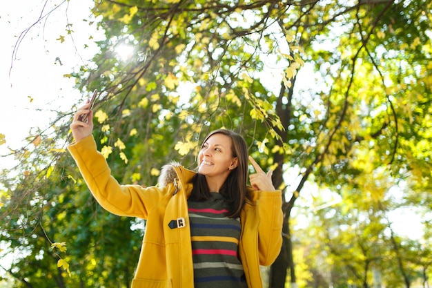 Die schöne fröhliche, fröhliche braunhaarige Frau in gelbem Mantel und gestreiftem Langarm macht Selfie auf dem Handy im Herbststadtpark an einem warmen Tag. Goldene Herbstblätter.