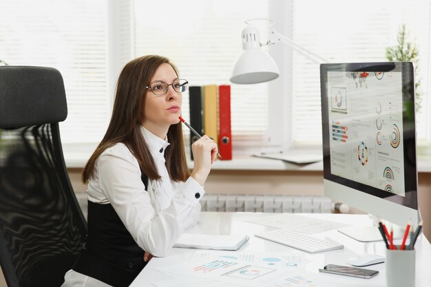 Die schöne Geschäftsfrau in Anzug und Brille, die am Computer mit Dokumenten im hellen Büro arbeitet und beiseite schaut