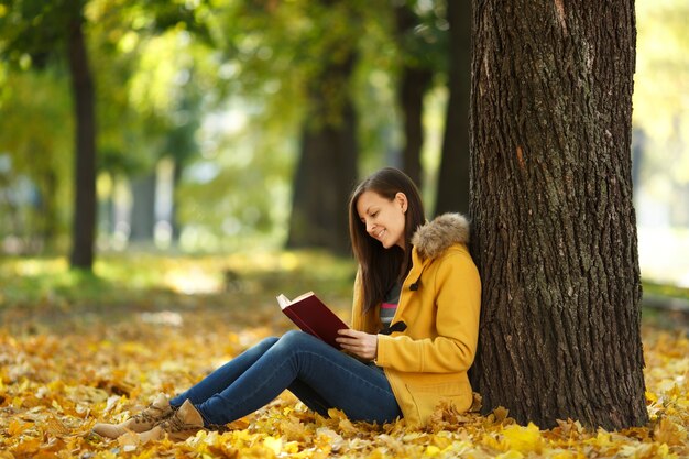 Die schöne glücklich lächelnde braunhaarige Frau in gelbem Mantel und Jeans, die an einem warmen Tag unter dem Ahornbaum mit einem roten Buch im Herbststadtpark sitzt. Goldene Herbstblätter. Lesekonzept