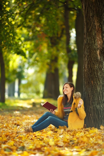 Die schöne glücklich lächelnde braunhaarige Frau in gelbem Mantel und Jeans, die an einem warmen Tag unter dem Ahornbaum mit einem roten Buch im Herbststadtpark sitzt. Goldene Herbstblätter. Lesekonzept