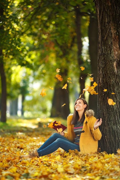 Die schöne glücklich lächelnde braunhaarige Frau in gelbem Mantel und Jeans, die an einem warmen Tag unter dem Ahornbaum mit einem roten Buch im Herbststadtpark sitzt. Goldene Herbstblätter. Lesekonzept