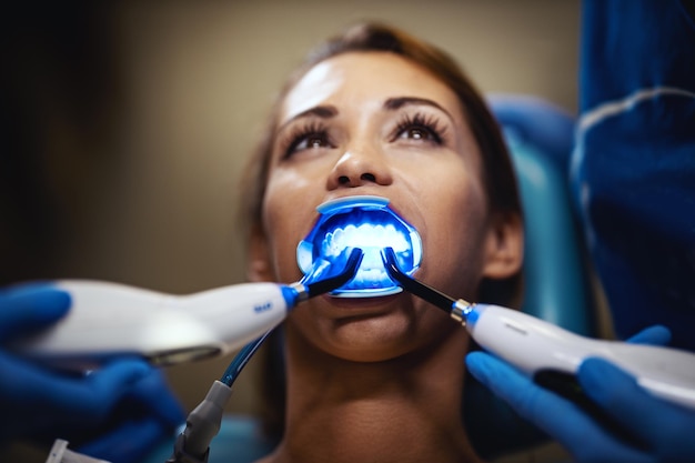 Die schöne junge Frau ist beim Zahnarzt. Sie sitzt im Zahnarztstuhl und der Zahnarzt setzt Zahnspangen auf ihre Zähne und setzt mit einer Infrarotlampe ästhetische, selbstausrichtende Lingualbrücken ein.