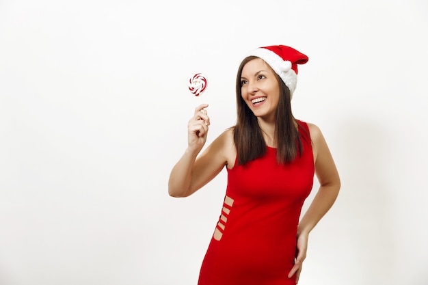 Die schöne kaukasische junge glückliche Frau mit gesunder Haut und charmantem Lächeln im roten Kleid und Weihnachtsmütze mit Lutscher auf weißem Hintergrund. Sankt-Mädchen isoliert. Neujahrsferien 2018 Konzept