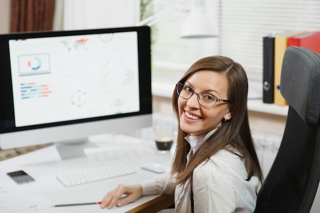 Die schöne lächelnde braunhaarige Geschäftsfrau in Anzug und Brille, die am Computer mit Dokumenten im hellen Büro arbeitet und in die Kamera schaut, gegen die Wand