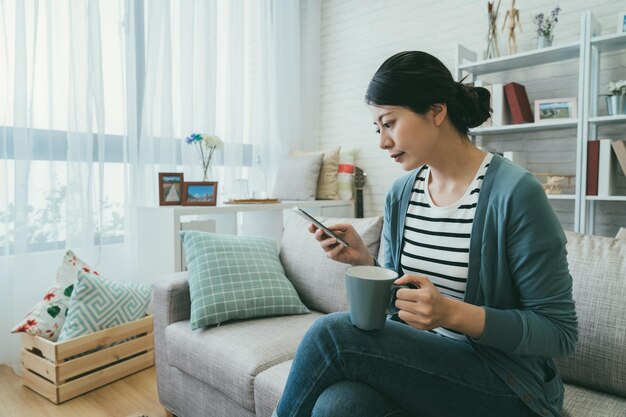 Die schöne taiwanesische Dame, die mit gefalteten Beinen auf dem Sofa sitzt, liest Online-Nachrichten auf dem Handy, während sie an einem ruhigen, sonnigen Morgen zu Hause Kaffee trinkt.