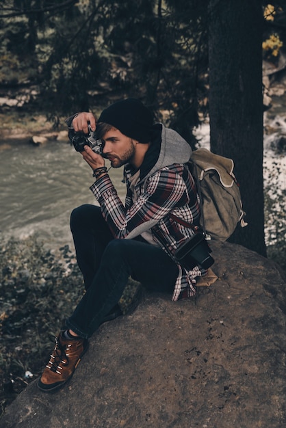 Die Schönheit einfangen. Junger moderner Mann mit Rucksack, der die Aussicht fotografiert, während er im Wald mit Fluss im Hintergrund sitzt