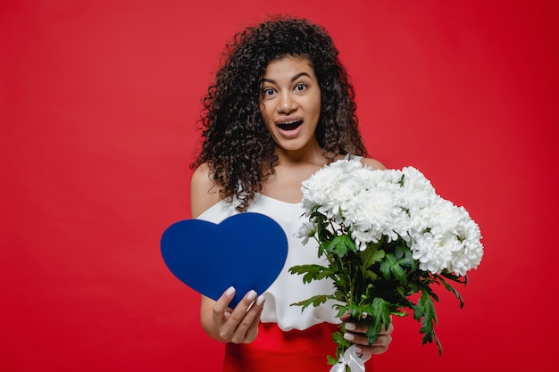 Die schwarze Frau, die mit blauem Herzen lächelt, formte Valentinsgrußkarte und Blumenstrauß von weißen Blumen