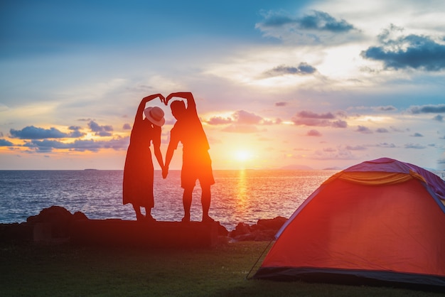 Die Silhouette des Hochzeitspaares, das Hände in einer Herzform am Meeresstrand bei Sonnenuntergang hält