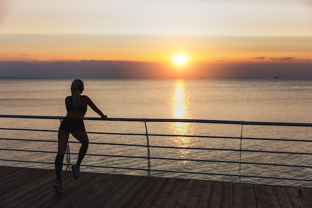 Die Silhouette eines jungen schönen athletischen Mädchens mit langen blonden Haaren in Kopfhörern, die bei Sonnenaufgang über dem Meer Musik hören und trainieren, Rückansicht