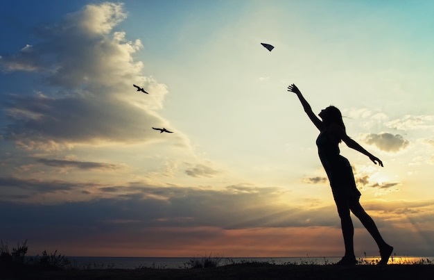 Die Silhouette eines Mädchens im Sommerkleid startet ein Papierflugzeug auf dem Hintergrund eines schönen Sonnenuntergangs