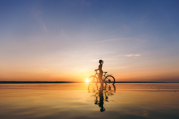 Die Silhouette eines sportlichen Mädchens in einem Anzug, das an einem warmen Sommertag bei Sonnenuntergang auf einem Fahrrad im Wasser sitzt. Fitness-Konzept.