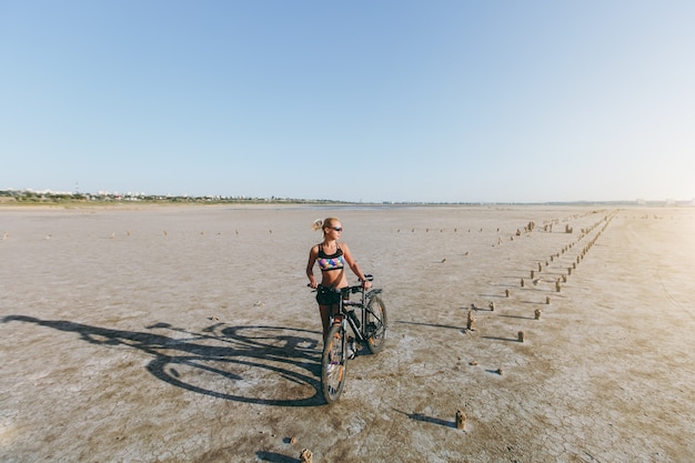 Die starke blonde Frau in einem mehrfarbigen Anzug und einer Sonnenbrille steht in einer Wüstengegend neben einem Fahrrad und schaut in die Sonne. Fitness-Konzept.