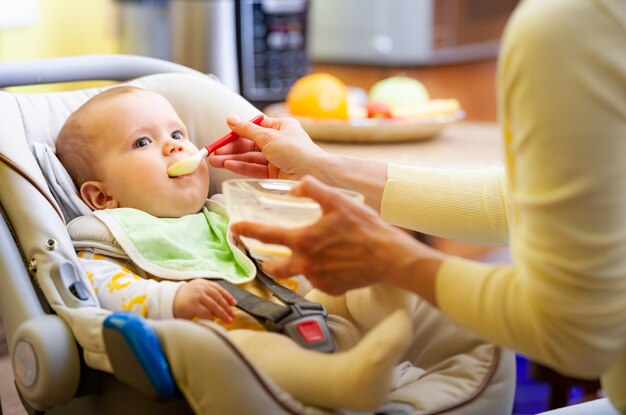 Die süße junge fürsorgliche kaukasische Mutter füttert ihre charmante kleine sechs Monate alte Tochter in einem gemütlichen Wohnzimmer.