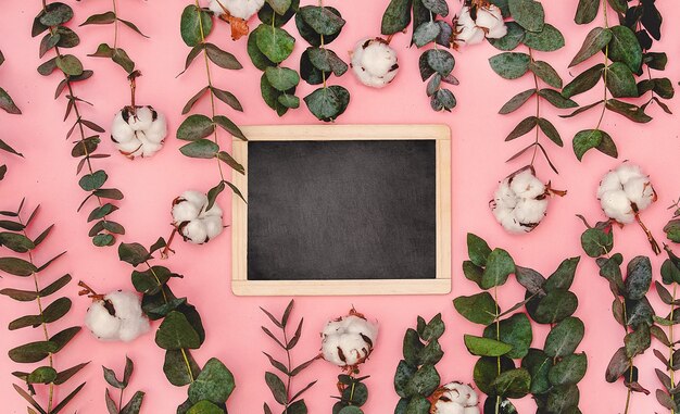 Die Tafel liegt auf dem rosa Tisch um die Blätter und Zweige von Eukalyptus und Baumwolle