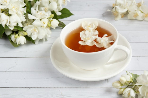 Die Tasse Tee, Honig aus einem Löffel in ein Glas gießend, blüht Jasmin auf einem hellen hölzernen Hintergrund.