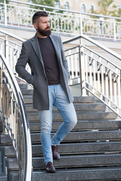 Die Treppe hinunter Stilvolles Freizeitoutfit Frühlingssaison Menswear und männliches Modekonzept Mann bärtiger Hipster stilvoller modischer Mantel oder Jacke Bequemes Outfit Hipster-Modemodell im Freien