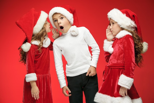 Die überraschten und glücklichen Jungen und Mädchen in Weihnachtsmannmützen mit Geschenkboxen im roten Studio