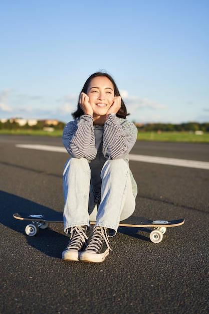 Die vertikale Aufnahme eines asiatischen Skatermädchens sitzt auf ihrem Skateboard und lächelt und genießt den sonnigen Tag beim Cruisen auf Lo