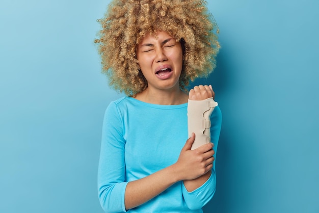 Die weinende, verärgerte, lockige junge Frau trägt einen elastischen Verband am Arm und muss in einer orthopädischen Klinik weiter behandelt werden. Sie leidet unter starken Schmerzen und wurde über blauem Hintergrund isoliert verletzt. Gesundheitsprobleme