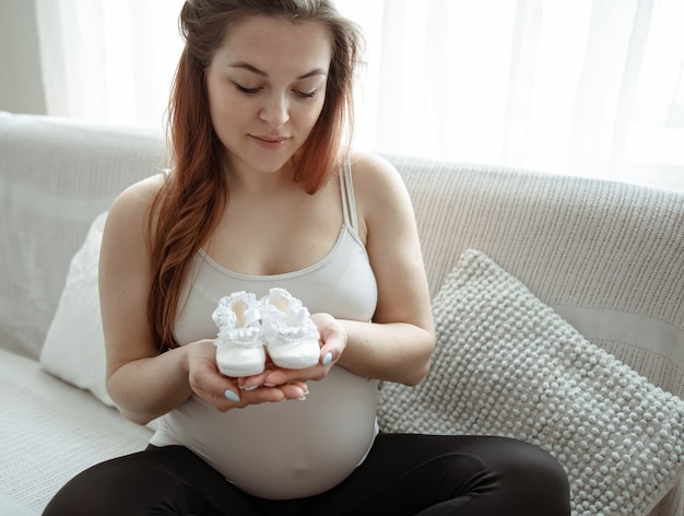 Die werdende Mutter mit dem dicken Bauch sitzt zu Hause mit Stiefeln für das Neugeborene auf der Couch.