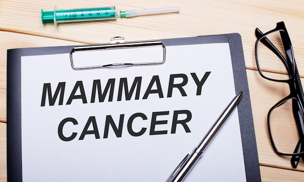 Die Worte MAMMARY CANCER stehen auf einem weißen Stück Papier neben einer schwarz umrandeten Brille, einem Stift und einer Spritze