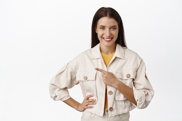 Diesen Weg. Porträt einer brünetten Frau in weißer, stylischer Jacke, Finger nach links zeigend und Verkaufspromo-Banner zeigend, Werbung, auf Weiß stehend.