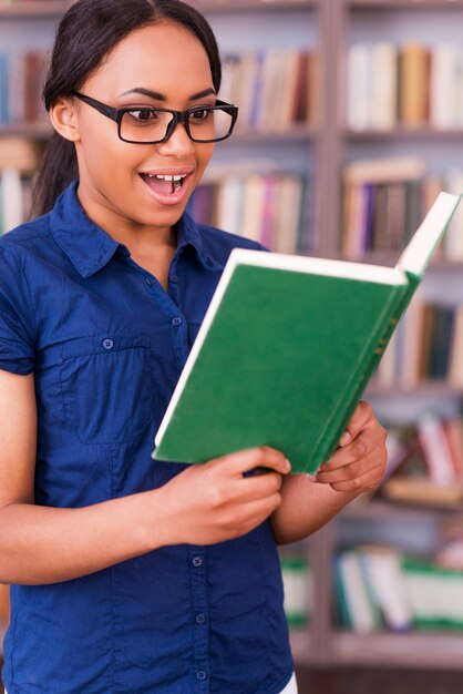 Dieses Buch ist so spannend! Überraschte afrikanische Studentin, die ein Buch liest und den Mund offen hält, während sie in der Bibliothek steht