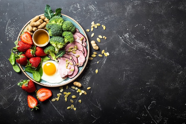 Dieta cetogénica baja en carbohidratos, vista superior, espacio para texto. Placa sobre fondo de piedra negra con alimentos cetogénicos: huevo, carne, aceite de oliva, brócoli, bayas, nueces, semillas. Grasas saludables, alimentación sana para bajar de peso