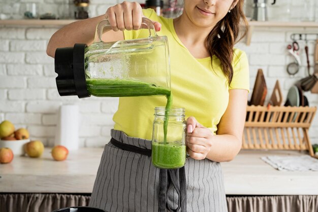 Dieta de alimentação saudável Jovem morena fazendo smoothie verde na cozinha de casa passando do liquidificador para a jarra