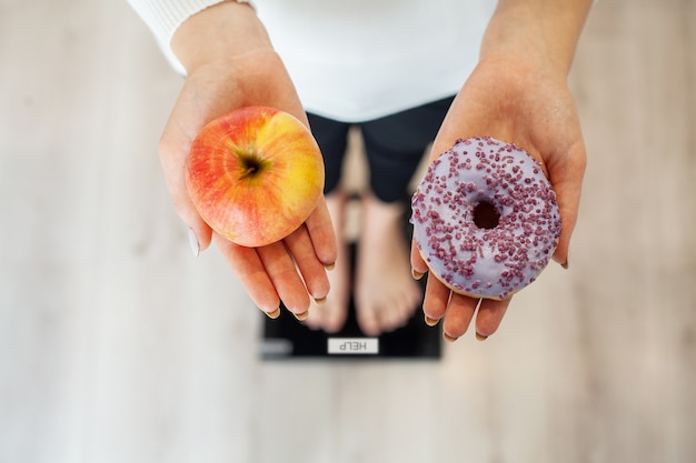 Dieta. Mujer que mide el peso corporal en la balanza Holding Donut y manzana. Los dulces son comida chatarra poco saludable. Dieta, alimentación saludable, estilo de vida. Pérdida de peso. Obesidad. Vista superior