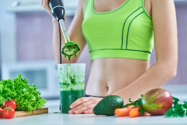 Dieta mujer en ropa deportiva prepara un batido verde con una batidora de mano en la cocina.