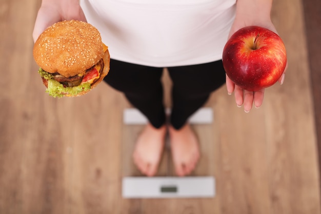 Foto dieta, mulher, medindo, peso corporal, ligado, pesando escala, segurando, hambúrguer, e, maçã