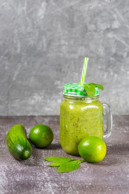 Dieta saudável suco de fruta fresca bebida com espinafre em uma jarra de vidro com um canudo sobre um fundo cinza de concreto rodeado por frutas e vegetais verdes.