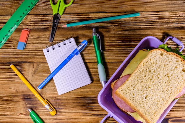 Diferentes artigos de papelaria e lancheira com sanduíches em uma mesa de madeira vista superior