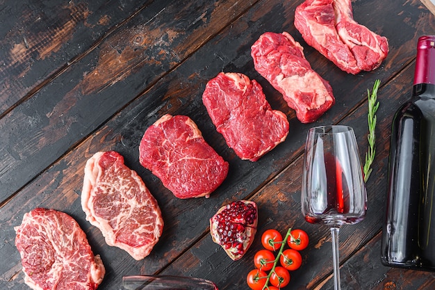 Diferentes bifes de carne crua com temperos e vinho tinto na garrafa e vidro na vista superior de pranchas de madeira escuras velhas com espaço para texto.