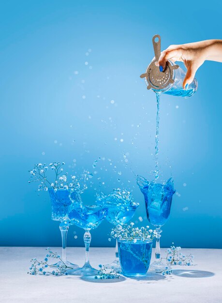 Diferentes taças de champanhe elegantes com coquetéis Blue Lagoon, a mão das mulheres está derramando coquetel do shaker para o copo coupe Splash splatter flores com gotas de água