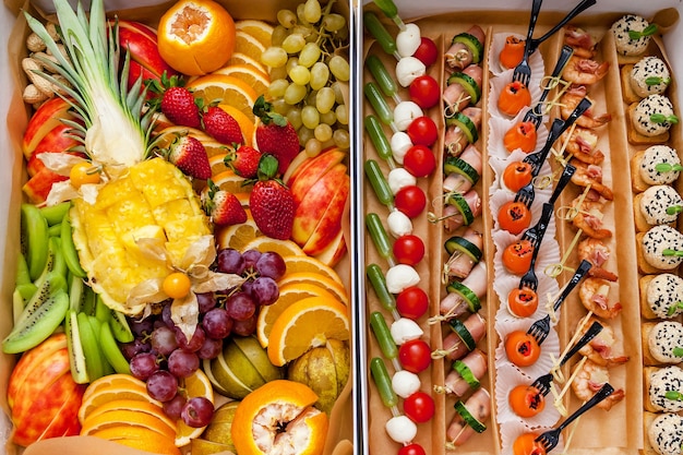 Diferentes tipos de canapés con pescado, verduras frescas y queso en una caja especial para pedido. Muchas frutas en rodajas como piña, naranjas, manzanas, kiwis, fresas.