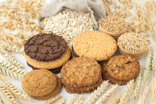 Diferentes tipos de galletas redondas, avena en bolsa, trigo y avena en cajas de madera. Espigas de trigo y avena sobre una mesa blanca