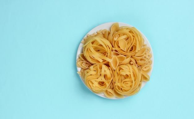 Diferentes tipos de pasta italiana cruda en placa sobre fondo azul.