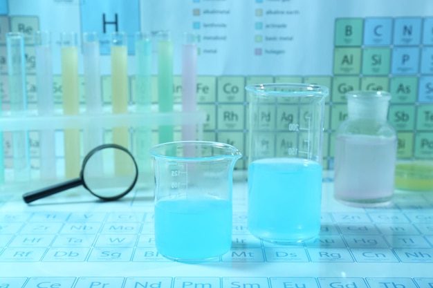Diferentes vasos de laboratorio con líquidos y lupa en la tabla periódica de elementos químicos Efecto de tono azul claro