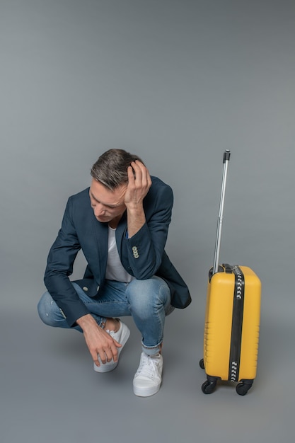 Foto dificuldade. homem triste com jaqueta jeans e tênis tocando a cabeça agachado perto da mala em antecipação