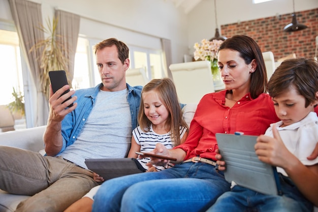 Digitale Ablenkungen Aufnahme einer jungen Familie, die zusammen auf ihrem Sofa im Wohnzimmer sitzt und von verschiedenen Medien und Geräten abgelenkt wird