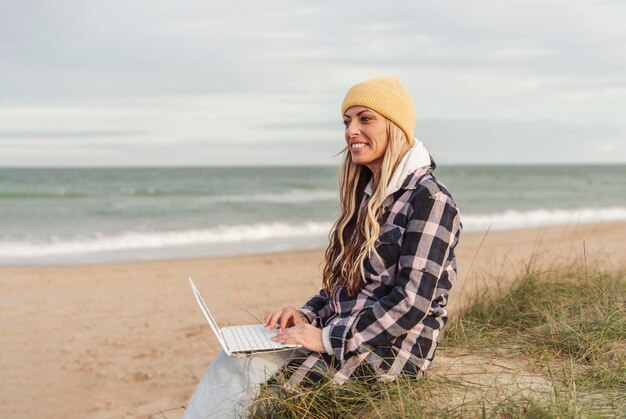 Digitale Frau des reisenden Nomaden im Winter oder im Herbst, die auf dem Strand unter Verwendung des Laptops sitzt