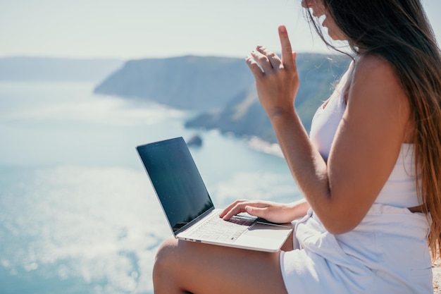 Digitale nomadenfrau mit hut eine geschäftsfrau mit laptop sitzt währenddessen auf den felsen am meer