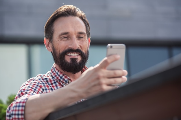 Digitale Stimmung. Freudiger gutaussehender Mann, der sein Telefon benutzt, während er auf der Bank sitzt