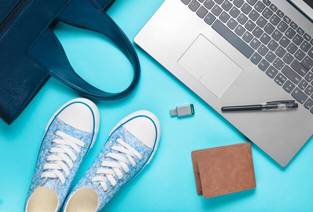 Digitale Technologie und modische Damenaccessoires auf blauem Hintergrund: Laptop, USB-Stick, Tasche, Brieftasche, Turnschuhe, Tasche. Draufsicht. Flach liegen.