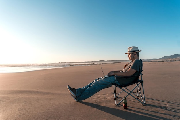 Digitaler Nomade sitzt auf Campingstuhl am Strand mit Laptop und macht unbeschwert Telearbeitx9