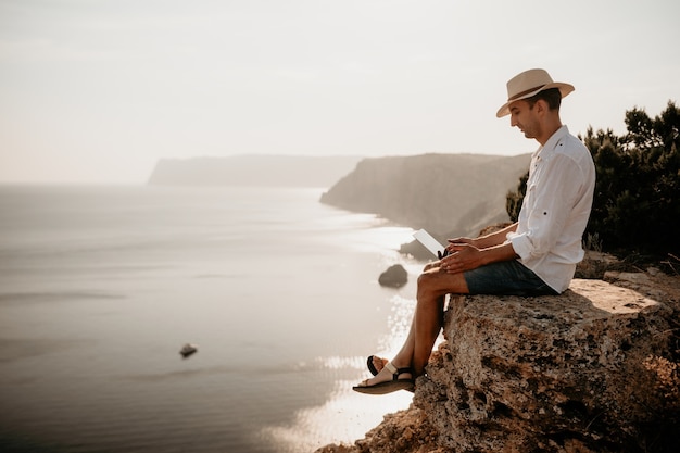 Digitaler Nomadenmann mit Hut ein Geschäftsmann mit Laptop sitzt bei Sonnenuntergang auf den Felsen am Meer